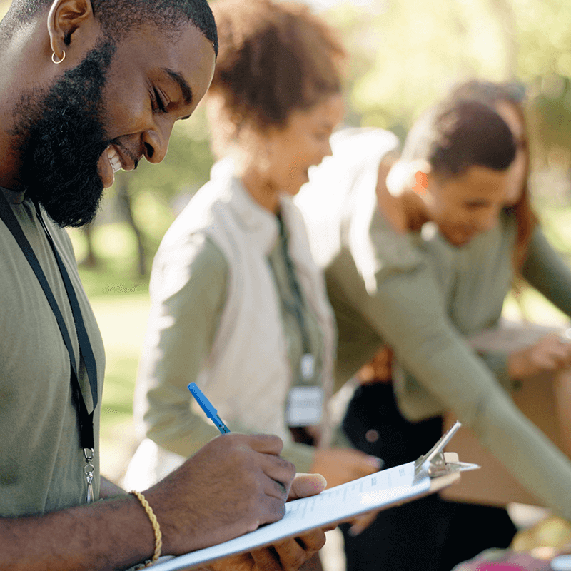 Un homme sourit au premier plan en prenant des notes devant d'autres personnes souriantes dans une forêt