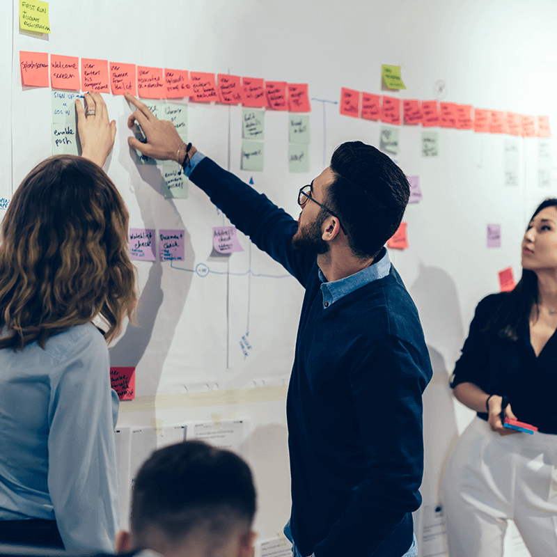 Un homme montre du doigt un tableau blanc plein de post-its devant 2 femmes